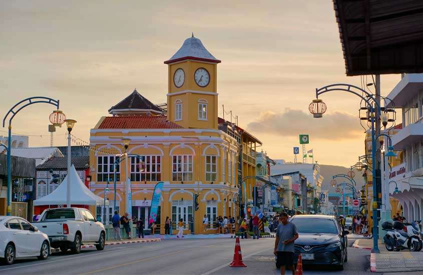 Durante o entardecer, rua de Phuket com construções coloridas, pessoas, carros e motos na rua