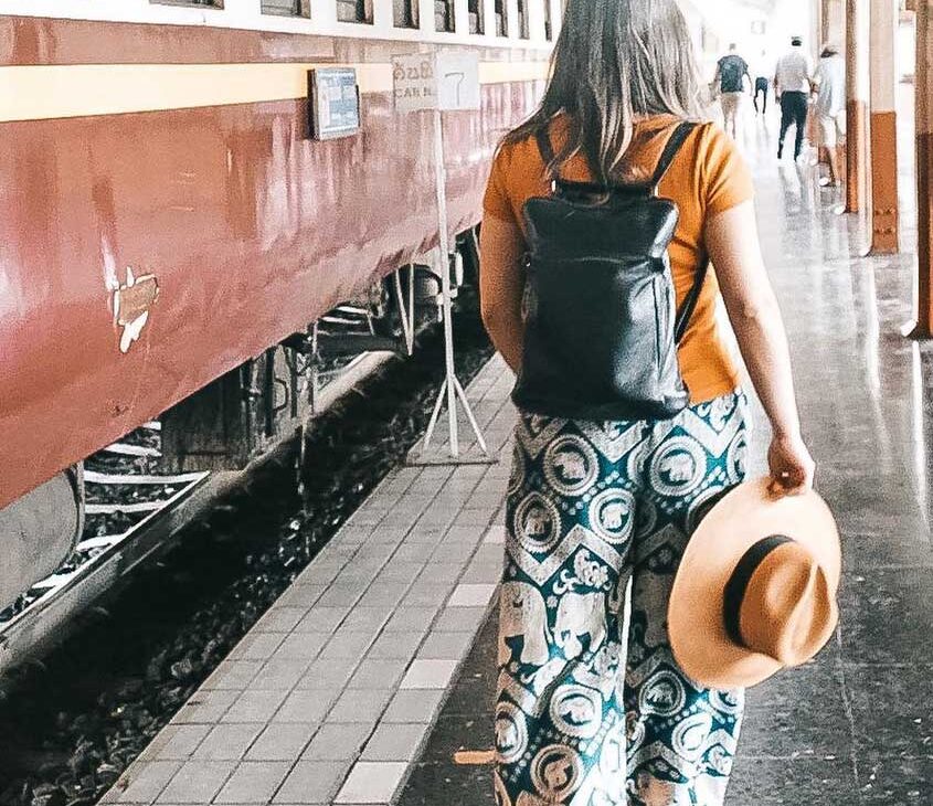 bárbara rocha, de costas, veste camisa laranja, calça estampada, carrega mochila preta nas costas, e segura chapéu, enquanto anda em estação de trem em chiang mai