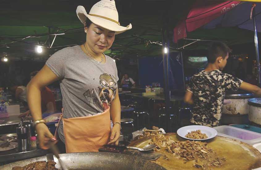 mulher, com chapéu de cowboy, camisa cinza e avental laranja, mexe em panela em barraca de comida de rua, durante a noite em um dos mercados em chiang mai