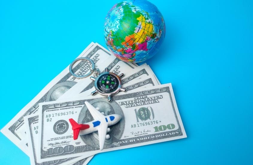 miniatura de avião, chaveiro de bússola e globo azul em cima de notas de dólares, em cima de mesa azul