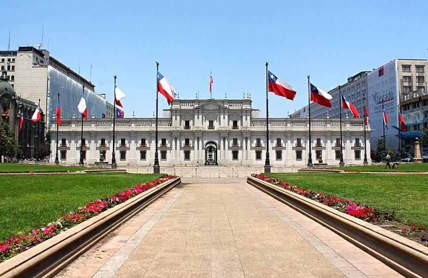 durante o dia, diversas bandeiras do chile hasteadas em frente a grande construção branca, onde funciona palacio de la moneda