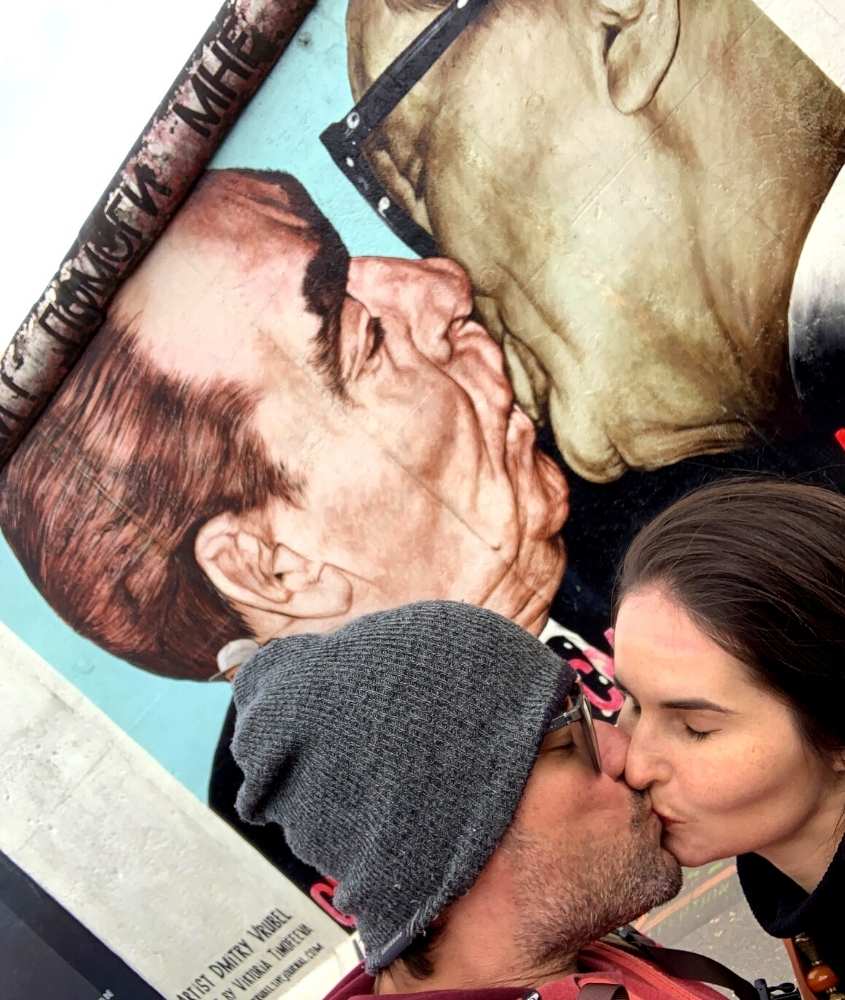 de touca cinza e óculos, vagner alcantelado beija bárbara rocha, em frente a pintura de muro de berlim, que exibe dois políticos se beijando, durante o dia