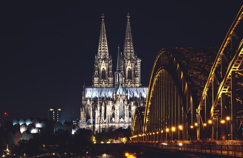ponte sobre o rio e ao fundo igreja iluminada, durante a noite em colônia, uma das melhores cidades da alemanha para visitar