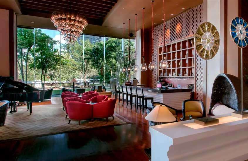Sala de hotel com poltronas, mesas, cadeiras, luminárias sofisticadas, bar e janela grande