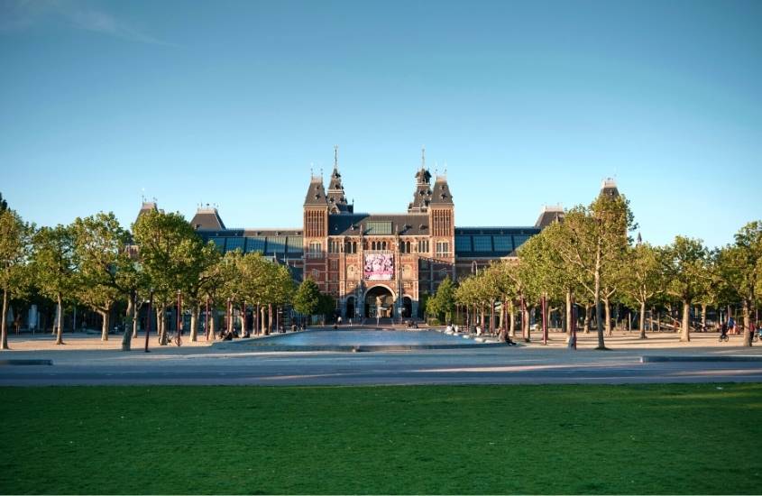 durante o dia, diversas árvores em frente a construção de tijolos vermelhos e telhado cinza em museumplein, um dos principais pontos turísticos de amsterdã