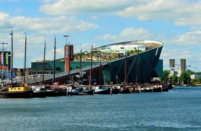 durante o dia, barcos em rio e ao fundo construção em formato de navio onde funciona um dos museus em amsterdã