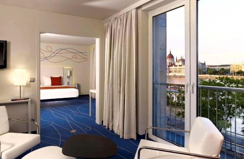 poltronas brancas, e ao fundo, cama de casal, em suite com vista para a cidade em um dos hotéis em budapeste no centro