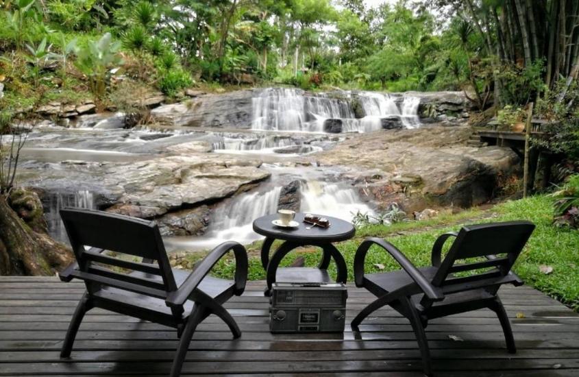 duas cadeiras marrons e pequena mesa redonda, em deck de madeira com vista para cachoeira, durante o dia