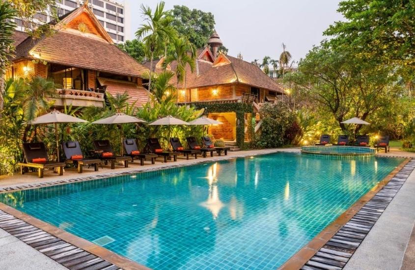 durante o dia, espreguiçadeiras e guarda-sóis em frente a grande piscina, cercada de árvores, em um hotel onde se hospedar em chiang mai