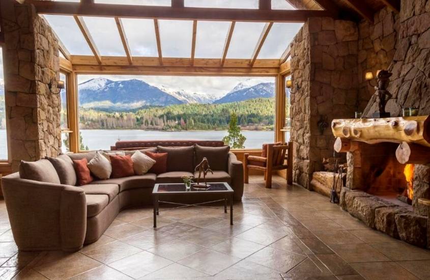 durante o dia, sofá em frente a lareira e ao fundo, ampla janela com vista para o lago em um dos melhores hoteis em bariloche para família