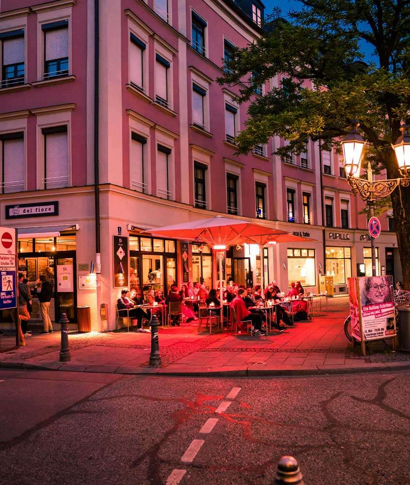 Durante o anoitecer, esquina com restaurante iluminado por luzes vermelhras, árvores ao lado e placas ao redor