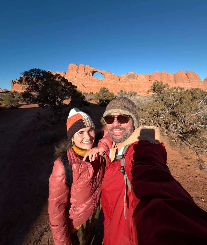 Em um dia de sol, Vagner Alcantelado e Bárbara Rocha Alcantelado, do blog melhores momentos da vida no Arches National Park com árvores ao redor e arcos atrás