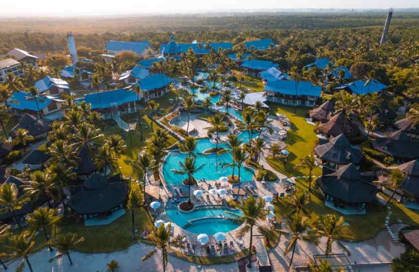 Em um dia de sol, visão aérea de um resort com piscinas, árvores, espreguiçadeiras e guarda-sois