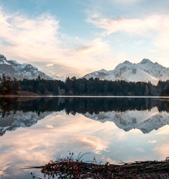 durante o dia, lago cercado de árvores e ao fundo montanhas cobertas de neve em bariloche