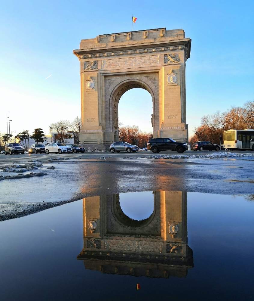 durante o vista, poça de água mostrando reflexo de construção de tijolos e mármore em formato de arco, e ao fundo, carros passando em frente ao arco