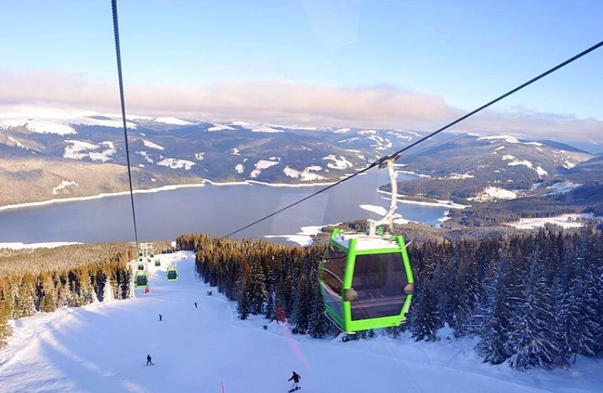 durante o dia, teleférico verde passando sobre montanha coberta de neve, onde há pessoas esquiando, ao fundo, lago