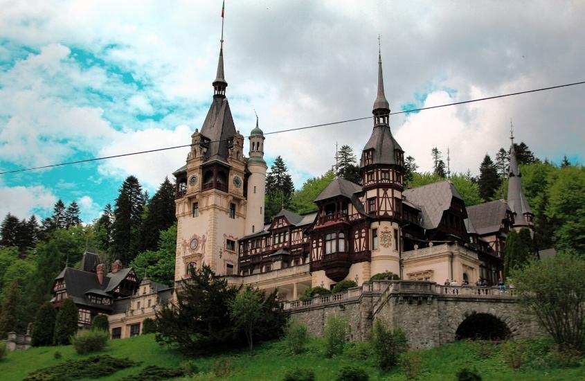 durante o dia, construção bege com telhados cinzas, onde funciona um dos castelos da romênia, rodeado por árvores