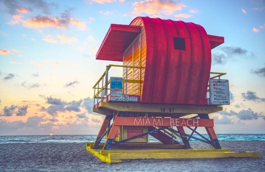 durante entardecer, cabana de salva-vidas vermelha e amarela onde há escrito 'miami beach', em areia de praia; ao fundo, mar