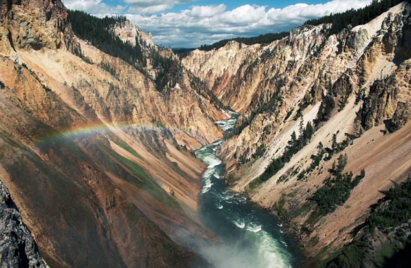 durante o dia, arco-íris passando entre grandes formações rochosas por onde passa um rio