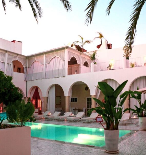 durante o dia, plantas, árvores e espreguiçadeiras ao redor de piscina, ao fundo, construção branca de dois andares onde funciona um dos riads em marrakech