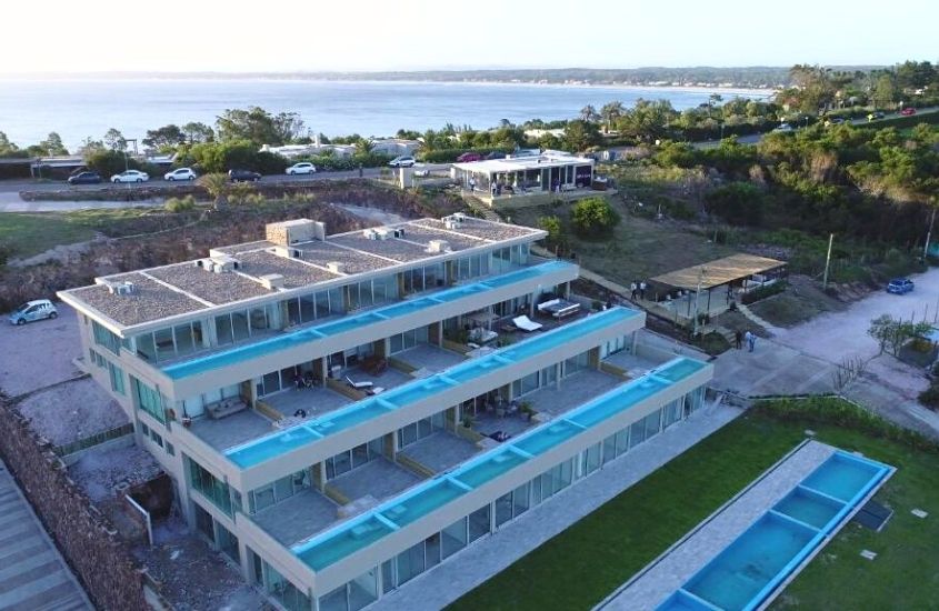 durante o dia, vista aérea de hotel com diversas piscinas em varanda de suítes, e ao fundo, mar