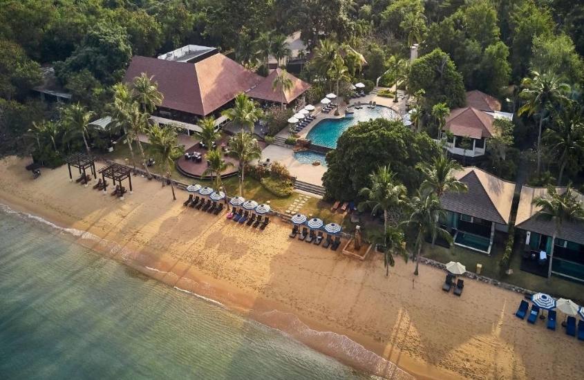 durante o dia, vista aérea, de mar, espreguiçadeiras em areia e ao fundo, complexo de villas de um resort na tailândia