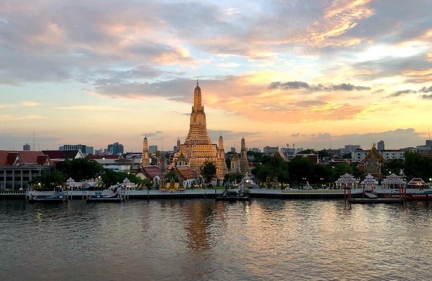 durante entardecer, barcos em rio e, ao fundo, templos e prédios em bangkok, destino para o roteiro tailândia