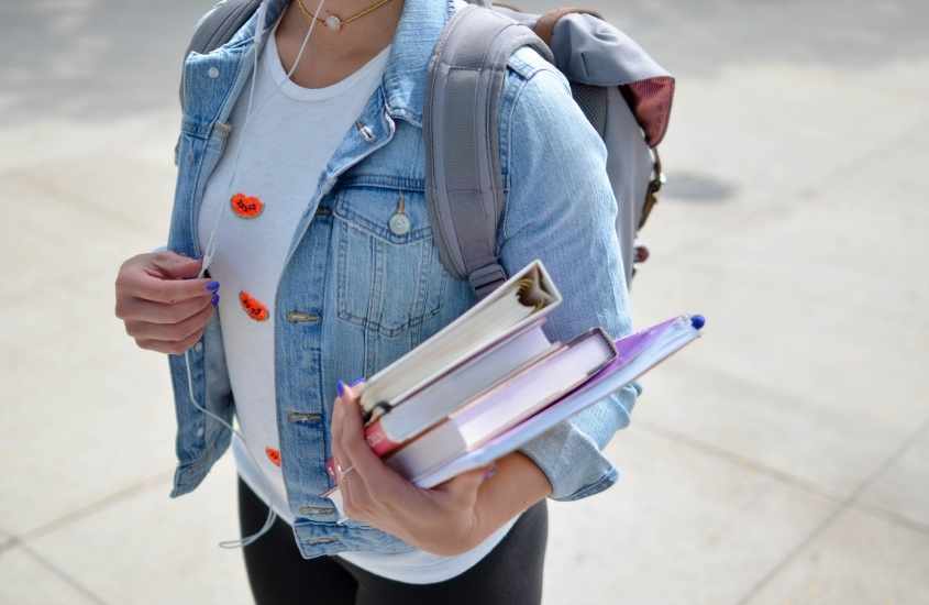 durante o dia, estudante vestida com blusa branca, jaqueta jeans e calça preta, carrega mochila cinza nas costas, e livros na mão
