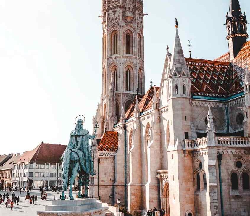 durante o dia, pessoas fazendo turismo em budapeste caminham em praça onde há estátuas e igrejas