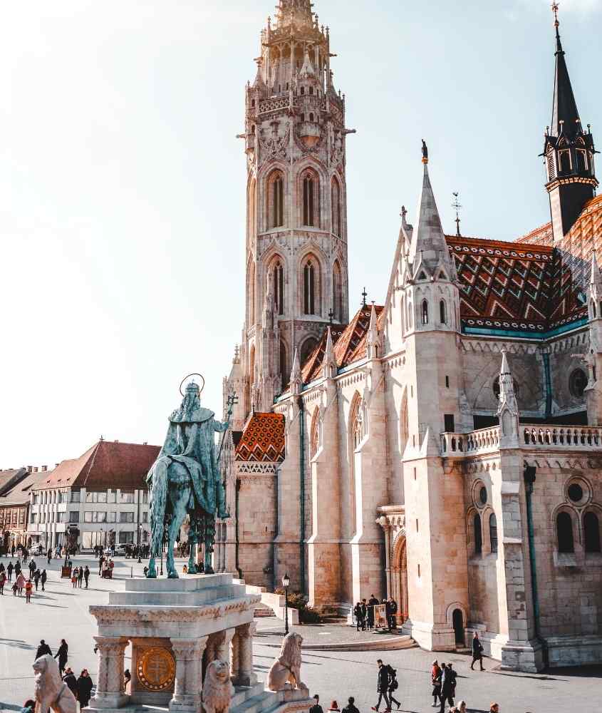 durante o dia, pessoas fazendo turismo em budapeste caminham em praça onde há estátuas e igrejas
