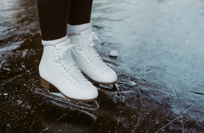pernas de pessoa vestindo calça preta e patins brancos, pisando em pista de gelo