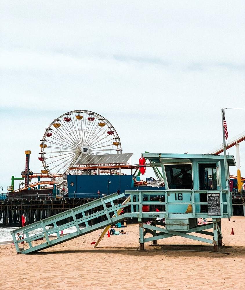 durante o dia, cabana azul sobre areia e, ao fundo, roda-gigante em cima de deck em uma das praias em los angeles