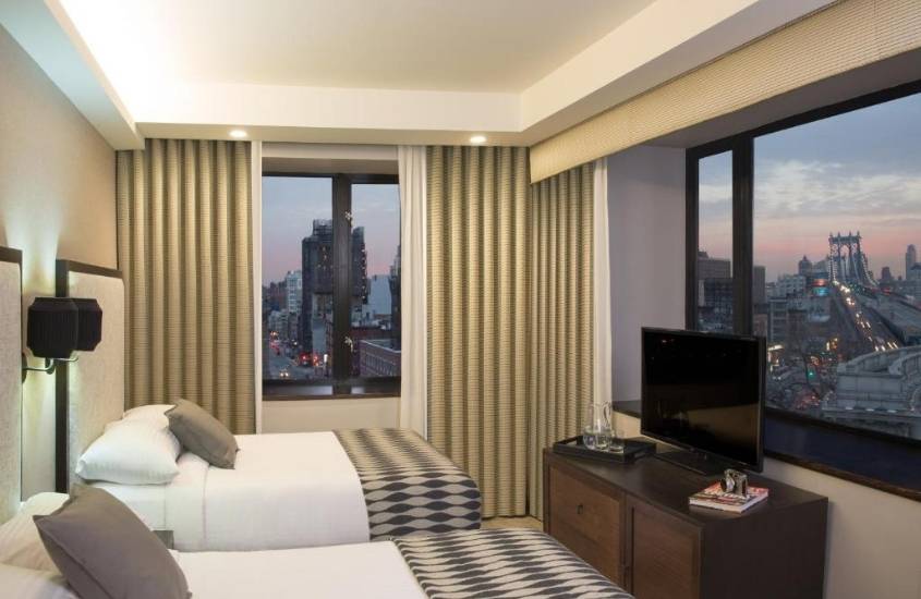 durante entardecer, camas de casal em frente a televisão, em suite com ampla janela com vista para a cidade
