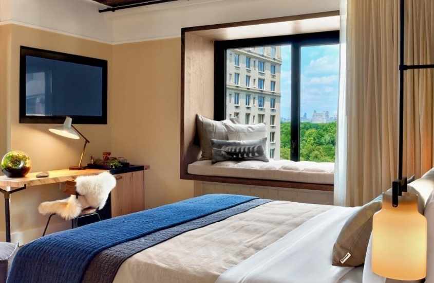 durante o dia, cama de casal, mesa de escritório e cadeira, em suíte com ampla janela com vista para a cidade em um dos hotéis em Nova York perto do Central Park