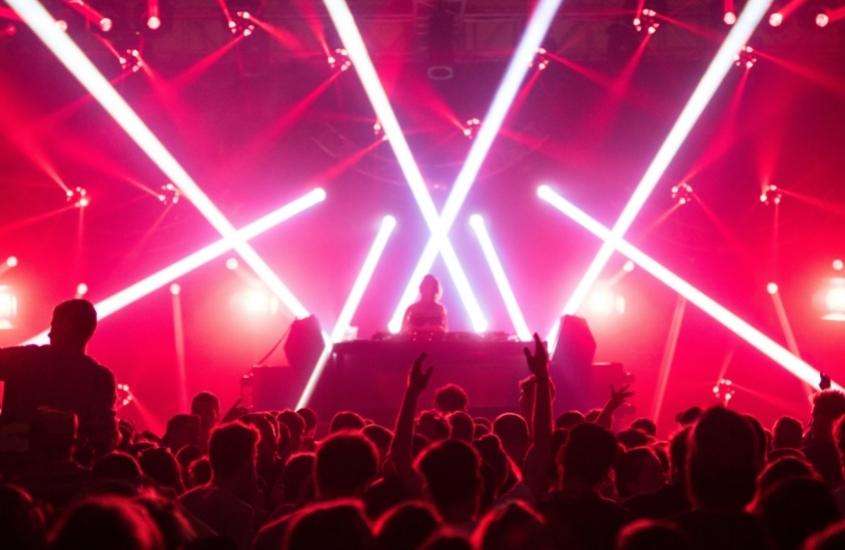 silhueta de pessoas de costa em frente a palco iluminado por luzes vermelhas, onde há DJ