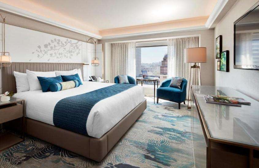 cama de casal e poltronas azuis em suite ampla, com vista para a cidade, durante o dia