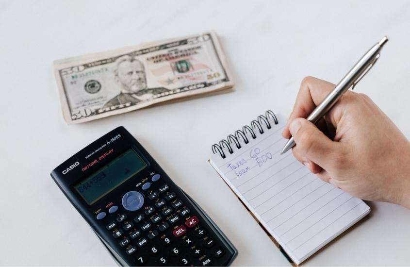 calculadora, notas de dólares e bloco de notas em cima de mesa branca para calcular preço de viagem para são francisco