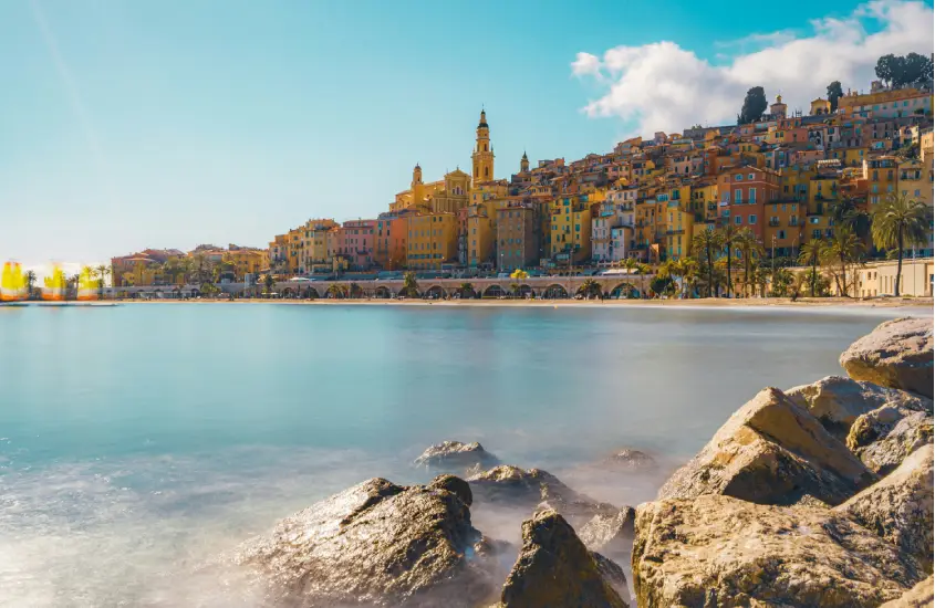 Em um dia ensolarado, paisagem de Menton com praia, árvores ao redor e construções coloridas