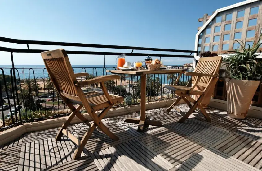 Em um dia de sol, varanda de um hotel com mesa, cadeiras, plantas decorativas, café da manhã e paisagem da Menton