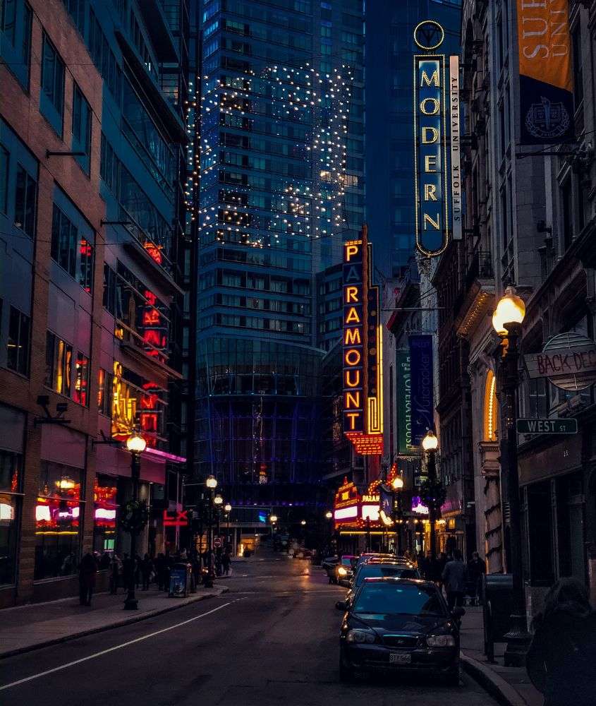 durante a noite, pessoas e carros passando em rua cheia de prédios e lojas com letreiros iluminados