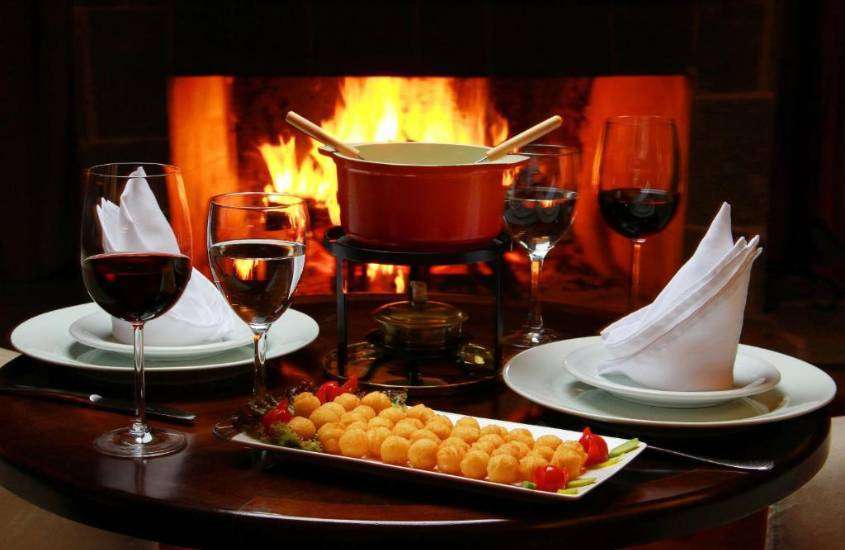 taça de vinho, salgados e fondue em mesa de madeira em frente a lareira, em hotel com ceia de réveillon em campos do jordão 2023