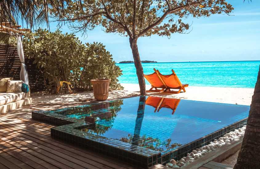 durante dia ensolarado, pequena piscina em deck ao livre e espreguiçadeiras laranjas em areia em frente ao mar