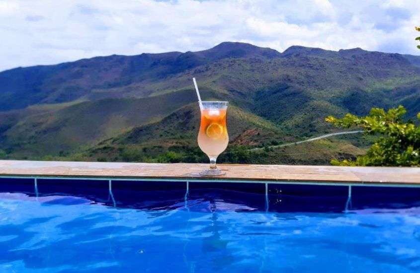 durante o dia, taça de vidro com drink laranja, em cima de beira de piscina com vista para as montanhas