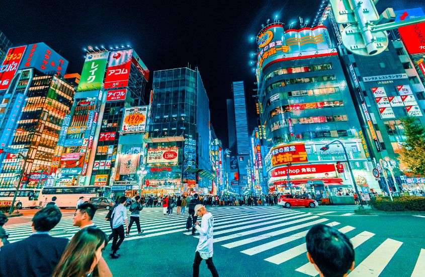 durante a noite, diversas pessoas atravessando rua movimentada de tokyo, cercada por prédios com letreiros iluminados