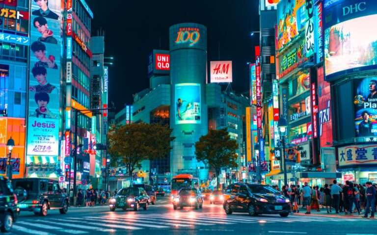 durante a noite, carros e pessoas passando em rua de um bairro de tokyo, cheio de prédios com letreiros iluminados