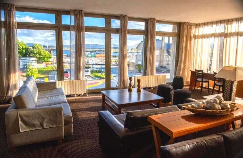durante dia ensolarado, sofás, mesa e cadeira em sala de estar de hotel em ushuaia com janelas panorâmicas com vista para a cidade