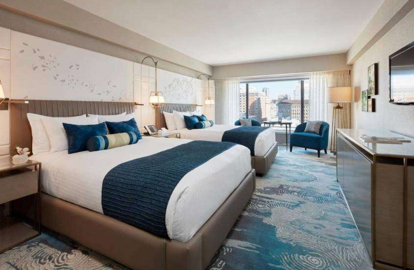 durante o dia, duas camas de casal e poltrona azul em quarto de hotel com vista para a cidade