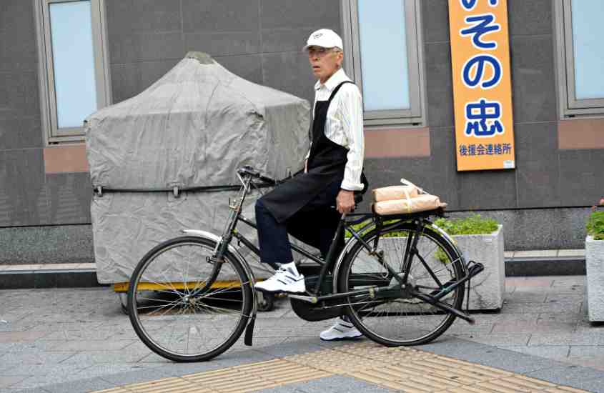 durante o dia, japonês idoso, com boné e camisa de manga longa brancos, avental e calça pretos e tênis branco, pedala em rua no japão