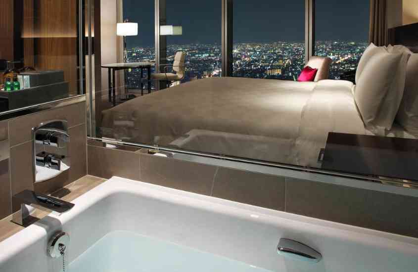 durante a noite, banheira de hidromassagem e cama de casal em suíte de um dos hotéis no japão com vista para prédios