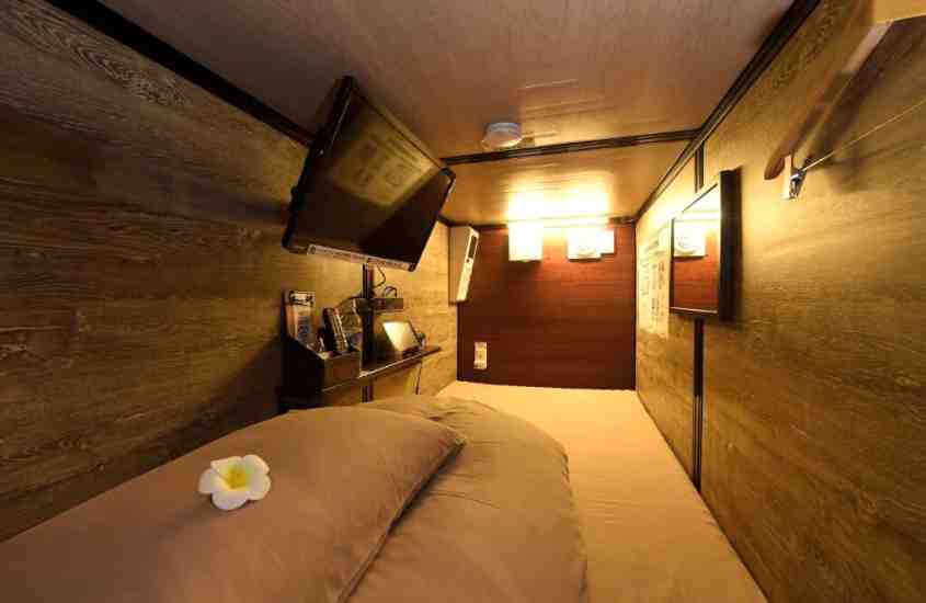 flor branca e amarela em cima de colchão cinza e TV em um hotel cápsula tokyo com paredes de madeira